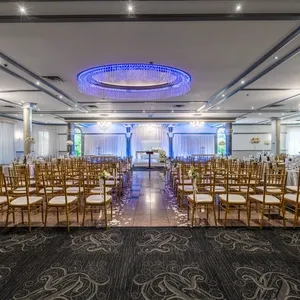 The image depicts an elegant wedding venue at Plaza Marina, showcasing a beautifully arranged ceremony setting. Rows of gold chiavari chairs are aligned neatly on either side of a glossy aisle, adorned with delicate floral arrangements. The backdrop features a tastefully illuminated stage, with draping and subtle lighting enhancing the romantic ambiance. The ceiling is adorned with an impressive chandelier and modern blue accent lighting, highlighting the spaciousness and grandeur of the venue. This setting exemplifies the sophisticated atmosphere offered by Plaza Marina for unforgettable wedding celebrations.