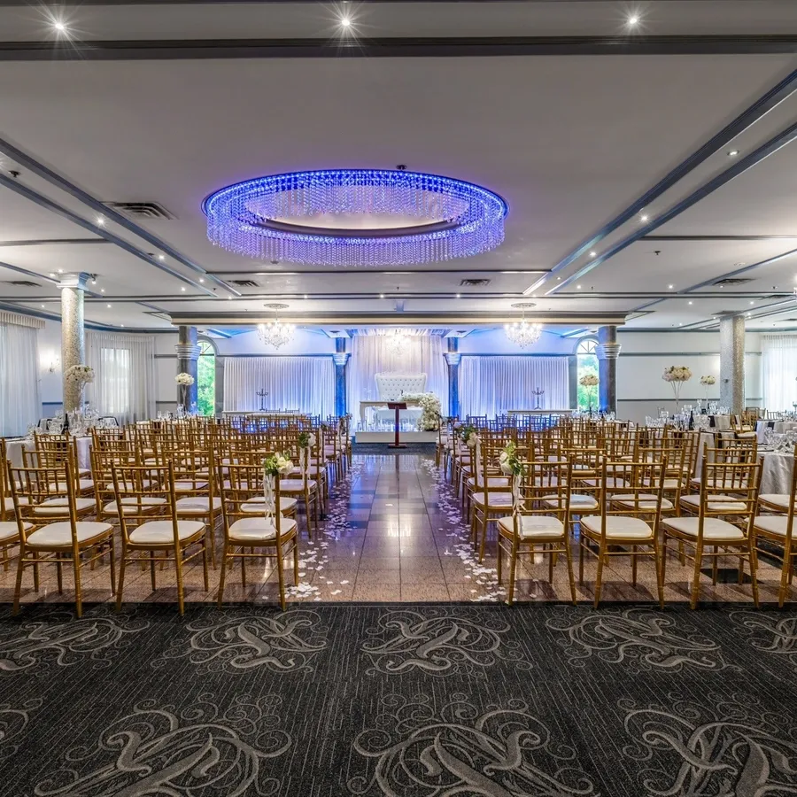 The image depicts an elegant wedding venue at Plaza Marina, showcasing a beautifully arranged ceremony setting. Rows of gold chiavari chairs are aligned neatly on either side of a glossy aisle, adorned with delicate floral arrangements. The backdrop features a tastefully illuminated stage, with draping and subtle lighting enhancing the romantic ambiance. The ceiling is adorned with an impressive chandelier and modern blue accent lighting, highlighting the spaciousness and grandeur of the venue. This setting exemplifies the sophisticated atmosphere offered by Plaza Marina for unforgettable wedding celebrations.