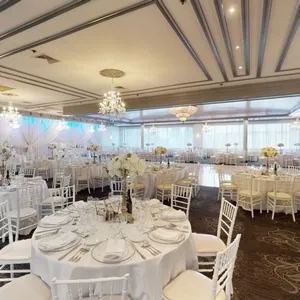In the image of Plaza Marina, a featured wedding venue on Mariage Elegant in Canada, a spacious and elegantly decorated banquet hall is shown. The venue is set for a wedding reception with round tables neatly arranged, each adorned with pristine white tablecloths and fine dinnerware. Elegant floral centerpieces grace each table, while the room is illuminated by ornate chandeliers that cast a warm glow over the setting. Delicate drapery hangs along the walls, adding a touch of sophistication to the atmosphere. The soft ambient lighting complements the classic white and pastel color scheme, creating a perfect backdrop for a memorable wedding celebration.