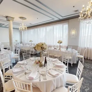 The image shows the elegant interior of Plaza Marina, a premier wedding venue listed under Mariage Elegant, Canada. The room features several round tables covered with pristine white tablecloths, each adorned with centerpieces of white flowers. Crystal chandeliers hang from the ceiling, providing a soft, ambient glow that complements the natural light streaming in through large, sheer-curtained windows. White chairs are neatly arranged around the tables, all set with fine china and glassware, creating a sophisticated atmosphere perfect for a wedding reception.