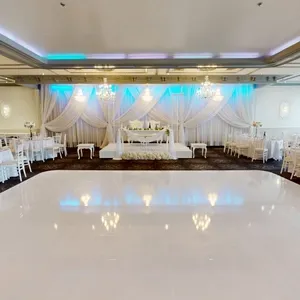 The image showcases an elegant wedding venue at Plaza Marina, a featured vendor on Mariage Elegant, Canada, under the Wedding Venues category. The spacious hall is adorned with luxurious chandeliers casting a warm glow over the room. White drapes elegantly frame a tastefully set head table at the center, complemented by blue ambient lighting. Round tables are neatly arranged around the polished dance floor, each set with white chairs and adorned with simple, yet stylish floral centerpieces, setting a sophisticated ambiance perfect for a wedding celebration.