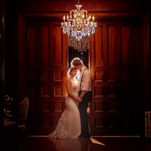 The image showcases a romantic wedding scene captured by Inhouse Studios Photography, a vendor specializing in wedding photography. In a dimly lit, elegant room with rich wooden paneling, a bride and groom share an intimate moment. They stand beneath a sparkling chandelier, creating a warm and inviting atmosphere. The bride is wearing a classic white gown with intricate lace details, and the groom is dressed in formal attire with suspenders. Their gentle embrace and the soft lighting highlight the emotion and elegance of the occasion.