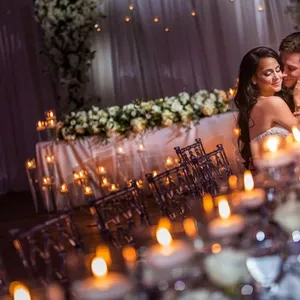 A romantic evening wedding reception photograph captured by Inhouse Studios Photography, exemplifying their expertise in wedding photography. The image features a newlywed couple lovingly embracing amidst a beautifully decorated setting. The atmospheric glow of numerous candlelit tables, elegantly arranged with florals, adds a warm and intimate ambiance. In the background, a stunning floral arch frames the couple, highlighting the festive environment suited for a wedding celebration.