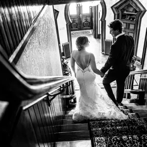 A bride and groom descend an ornate wooden staircase in an elegant, historic venue. The bride is wearing a stunning lace wedding dress with a long train, while the groom is in a classic dark suit. They are captured from behind, highlighting the intricate details of the bride's gown. Soft light filters through a window, creating a warm, romantic ambiance. This image showcases the sophisticated and artistic style of Inhouse Studios Photography, a vendor specializing in wedding photography, exemplifying their ability to capture intimate, beautiful moments for weddings in Canada.