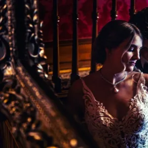 Inhouse Studios Photography captures an intimate moment of a bride and groom in a dimly lit staircase setting. The bride is wearing an intricate lace wedding gown with a delicate pearl necklace that complements her elegant look. The groom is dressed in a classic black tuxedo, gently leaning towards the bride. The setting features a luxurious wooden banister and rich, red wallpaper that add a romantic and timeless atmosphere to the scene. This image highlights the wedding photography expertise of Inhouse Studios Photography, showcasing a blend of style and emotion perfect for couples planning their special day with Mariage Elegant in Canada.