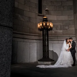 In this elegant wedding photography image by Inhouse Studios Photography, a bride and groom are seen embracing under a grand, ornate lamp post in a sophisticated stone setting, highlighting the romantic and intimate mood of their special day. The bride is wearing a stunning, white lace gown with a flowing veil cascading to the floor, while the groom is in a classic dark suit, perfectly complementing the luxurious and timeless backdrop. This photo showcases the artistry of wedding photography, capturing a tender moment against the dramatic architecture, ideal for those seeking timeless elegance in their wedding memories.