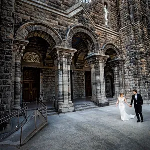 A bride and groom hold hands as they walk in front of a grand stone cathedral with intricate archways and ornate columns, captured by Inhouse Studios Photography. The bride wears an elegant white gown, while the groom is dressed in a sharp black tuxedo. This wedding photography showcases a serene and timeless moment, highlighting the couple's joyous occasion in a historic setting. The image is part of the wedding photography category on Mariage Elegant, Canada, featuring the artistry of Inhouse Studios Photography.