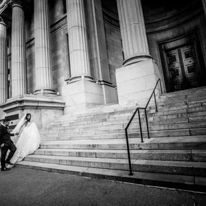 In this black and white image by Inhouse Studios Photography, a newlywed couple is captured in the wedding photography category for Mariage Elegant. The bride, wearing a flowing white gown with a long train, descends the grand stone steps of an impressive, column-lined building. Her groom, in a dark suit, gently holds her hand. The architectural detail of the venue provides a classic and timeless backdrop, enhancing the elegance of the moment. The scene conveys a sense of romance and celebration, ideal for a wedding photography portfolio.