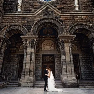 A bride and groom stand intimately in front of a grand, historic stone church facade, showcasing intricate architecture with tall arches and detailed carvings. The bride wears an elegant white gown with a flowing train, while the groom is dressed in a classic black suit. They are positioned in the center, holding hands and gazing at each other, capturing a moment of affection. The sunlight casts gentle shadows, highlighting the textures of the church's stonework. This image, taken by Inhouse Studios Photography, beautifully encapsulates the essence of wedding photography, emphasizing romance and architectural elegance.
