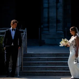 The image shows a bride and groom in an elegant, dimly lit outdoor setting. The bride is wearing a beautiful white lace wedding gown and holding a bouquet of white flowers, while the groom stands opposite her in a classic black tuxedo with a white shirt and black bow tie. They are positioned in front of a grand stone staircase, which adds a touch of sophistication to the scene. This image captures a romantic moment, perfect for showcasing the skills of Inhouse Studios Photography, a vendor specializing in wedding photography within the Mariage Elegant listings in Canada.
