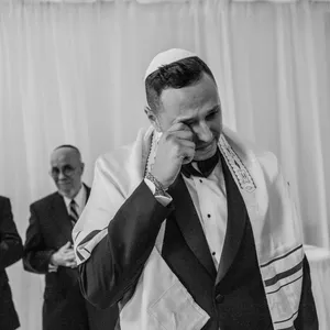 Inhouse Studios Photography captures an emotional moment in the wedding ceremony, focusing on a groom standing in front of a white draped backdrop. The groom is dressed in a formal black tuxedo with a white kippah and tallit, wiping away tears of joy. In the background, several guests, including two men in suits and a woman in a sequined dress, watch the scene with expressions of happiness. This image, showcasing wedding photography, highlights the heartfelt emotions present during a marriage celebration, perfectly aligning with the services of Inhouse Studios Photography under the wedding photography category on Mariage Elegant, Canada.