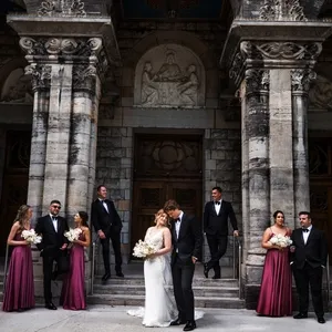 This image from Inhouse Studios Photography, highlighting their expertise in wedding photography, captures a sophisticated wedding party posed outside a grand, historic stone building with ornate columns. The couple stands gracefully at the center, with the bride holding a beautiful bouquet, while the bridal party, dressed in elegant attire with the bridesmaids in matching burgundy dresses, complements the scene. The mood is romantic and timeless, aligning with the elegant wedding theme.