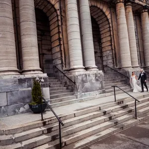 A newlywed couple walks hand-in-hand down the grand stone steps of a historic, columned building, evoking a timeless elegance. The bride, dressed in a traditional white gown, carries a bouquet, while the groom in a classic black suit, complements her grace. This captivating image by Inhouse Studios Photography falls under the Wedding Photography category on Mariage Elegant, Canada, showcasing their ability to capture romantic and sophisticated moments in iconic settings. The architecture's grandeur adds a majestic backdrop to the couple's joyful journey.