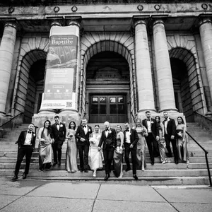 A wedding party poses on the steps of a grand, historic building with large stone columns in the background. The group includes bridesmaids in elegant dresses and groomsmen in formal suits, all smiling and standing close together. This image showcases the work of Inhouse Studios Photography, capturing a joyful moment in wedding photography. The scene conveys a sense of timeless celebration, with the architectural details adding a majestic backdrop to the occasion. The photo is suitable for the vendor listing on Mariage Elegant, Canada.