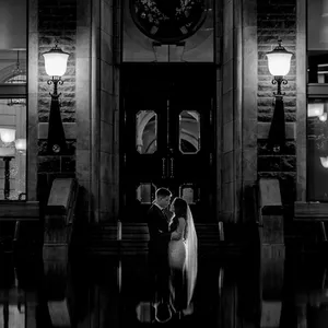 A black and white photograph showcases a bride and groom embracing in front of an elegant building, beautifully captured by Inhouse Studios Photography, a vendor in the Wedding Photography category. The couple stands illuminated under glowing street lamps that frame the entrance, creating a romantic and timeless atmosphere. The reflection of the couple and the ornate architecture in a still pool of water below adds a dramatic and artistic effect, perfect for a memorable wedding moment.