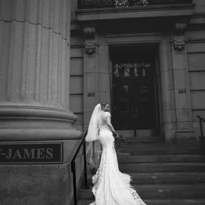 This black and white photograph by Inhouse Studios Photography captures a bride elegantly posed on the steps of a grand building, highlighting the wedding photography category. The bride is wearing a stunning, intricate lace gown with a long train and a veil, creating a timeless and sophisticated look. The setting features tall columns and architectural details, adding to the grandeur of the scene. The image reflects the elegance and beauty that Mariage Elegant, Canada's wedding planning service, aims to promote through its vendor listings.