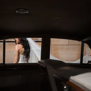 A bride in a white, strapless lace wedding gown is captured walking alongside a vintage car, her long veil being elegantly carried by a person in a tuxedo. This striking scene, photographed by Inhouse Studios Photography, exemplifies their expertise in capturing intimate and stylish moments in the wedding photography category. The image highlights the elegance and sophistication expected in a wedding setting, showcased against a classic backdrop.