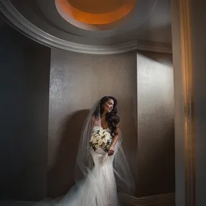 The image features a bride elegantly standing under a soft spotlight in a beautifully curved alcove with a decorative round ceiling, captured by Inhouse Studios Photography. She is wearing a flowing white wedding gown with a long, delicate train and holding a bouquet of pale roses, highlighting the category of wedding photography. Her veil drapes gracefully over her shoulders, enhancing the romantic atmosphere. The setting exudes an intimate and serene ambiance, perfectly suited for Mariage Elegant's wedding planning and vendor listings in Canada.
