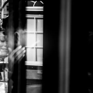 This black-and-white image by Inhouse Studios Photography captures an intimate and emotional moment in the wedding context. The photograph shows a bride, slightly obscured and reflected in a windowpane, creating an artistic and dreamy effect. Her expression suggests deep contemplation, complemented by the soft light and shadow play across her face. The background features a window with geometric patterns, adding a touch of elegance and sophistication to the composition. This image wonderfully encapsulates the essence of wedding photography, showcasing both artistic style and emotional depth.
