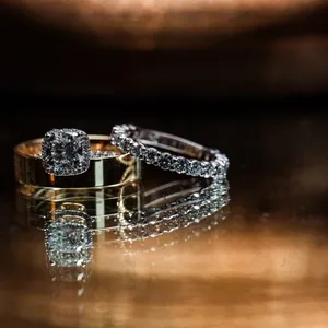 The image showcases two beautifully crafted wedding rings placed elegantly on a reflective surface. One ring features a stunning diamond engagement ring with a halo setting, while the other is a classic gold band. The background is softly blurred, creating a warm, romantic ambiance. This captivating photograph is captured by Inhouse Studios Photography, a vendor specializing in wedding photography, and is perfect for conveying elegance and commitment for Mariage Elegant, Canada.