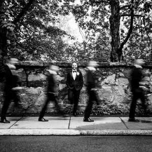 The image features a groom in formal attire, standing confidently against a rustic stone wall, surrounded by a lush, tree-filled background. He is captured in a serene pose while groomsmen, dressed in similar suits, briskly walk in front of him, creating a striking motion blur effect. This black-and-white photograph, presented by Inhouse Studios Photography, showcases their expertise in capturing unique and dynamic wedding moments. The photo highlights the artistry of wedding photography, emphasizing both the stillness and motion of a wedding day, suitable for Mariage Elegant in Canada.