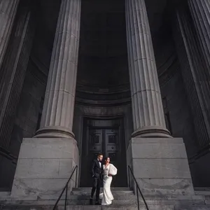 A newlywed couple poses on the steps of a grand building with towering stone columns, showcasing Inhouse Studios Photography's expertise in wedding photography. The bride is elegantly dressed in a white gown, while the groom is in a dark suit. The image captures the dramatic architecture and classic romance, perfect for a wedding setting. This photo exemplifies the high-quality offerings of Inhouse Studios Photography, listed on Mariage Elegant, Canada.