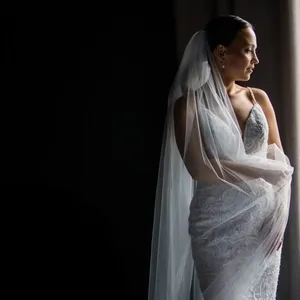 A bride stands gracefully in a softly lit room, captured by Inhouse Studios Photography under the wedding photography category. She is wearing a delicate, lace wedding gown with a fitted bodice and a flowing veil over her shoulders. The bride's hair is elegantly styled back, highlighting her serene profile as she gazes to the right. The image showcases the elegance and attention to detail typical of a wedding day, reflecting the romantic atmosphere and professional expertise of Inhouse Studios Photography.