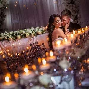 A bride and groom share an intimate moment at their elegantly decorated wedding reception, captured by Inhouse Studios Photography in the wedding photography category. The bride is wearing a strapless gown and the groom is in a dark suit. They are seated closely together, surrounded by softly glowing candles and a beautifully set table with floral arrangements, creating a romantic atmosphere. The background showcases a draped fabric and more flowers, contributing to the overall sophisticated and elegant ambiance.