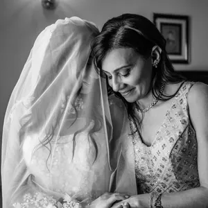 Inhouse Studios Photography captures an intimate black and white wedding moment under the wedding photography category. The image shows a bride in a delicate veil and intricate floral gown gently leaning towards another woman in an elegant sleeveless dress adorned with subtle embellishments. Both women share a tender smile, exuding warmth and connection in a softly lit room, perfect for this serene moment. The background features a framed picture on the wall, suggesting a cozy, personal setting often sought in weddings.