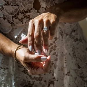A close-up photograph by Inhouse Studios Photography in the wedding photography category shows a bride's hands delicately clasped together. The focus is on her elegant, large diamond engagement ring that sparkles in the light. She wears a beautiful lace wedding dress, highlighting intricate floral patterns that add a classic, romantic touch. Her nails are neatly manicured with a soft, pale pink polish, complementing the overall elegant bridal look.