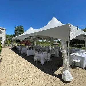 The image shows an elegantly set-up outdoor wedding venue at Plaza Rive Sud, categorized under Wedding Venues for Mariage Elegant in Canada. A large white tent is centrally placed on a spacious, paved patio surrounded by lush greenery and gardens. Inside the tent, numerous white-clothed tables with neatly arranged white chairs are prepared for a wedding reception. The bright blue sky and beautifully manicured landscape enhance the romantic and tasteful ambiance, making it an ideal location for a sophisticated outdoor wedding celebration.
