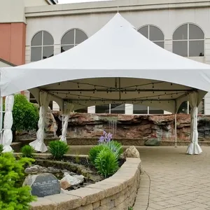 The image features Plaza Rive Sud, a premier wedding venue located in Canada, showcasing a spacious outdoor area ideal for weddings. A large white event tent stands prominently in the center, providing a shaded space for guests. The tent is elegantly draped and adorned with string lights, creating a romantic ambiance. Surrounding the tent is a beautifully landscaped area with lush greenery, blooming flowers, and a tranquil waterfall feature set in a stone wall, enhancing the picturesque setting for a memorable wedding ceremony or reception.