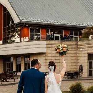 A bride and groom walk hand in hand toward the elegant Plaza Rive Sud, a premier wedding venue in Canada. The bride is wearing a beautiful lace gown and holding a vibrant bouquet of flowers high in the air. The groom is dressed in a stylish blue suit. The venue features modern architecture with large glass windows and a spacious outdoor area, perfect for wedding ceremonies and receptions. The setting reflects a joyful celebration, highlighting the sophisticated charm of Plaza Rive Sud within the wedding venues category on the Mariage Elegant site.