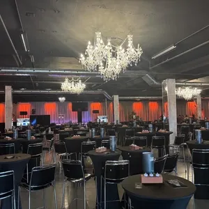 The image showcases the interior of Plaza Rive Sud, a distinguished wedding venue in Canada listed under the wedding venues category on Mariage Elegant. The spacious room features elegant black round tables and tall chairs, set with stylish centerpieces and modern tableware, creating a sophisticated atmosphere perfect for a wedding reception. The dimly lit environment is enhanced by luxurious crystal chandeliers hanging from the ceiling, casting a warm glow that complements the vibrant red curtains adorning the walls. This venue offers a chic and modern setting for an unforgettable wedding celebration.