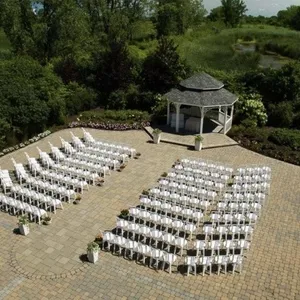 The image shows an outdoor wedding venue at Plaza Rive Sud, categorized under wedding venues. It features a beautifully arranged setting with rows of white chairs on a spacious stone-paved area. At the center is a charming white gazebo, ideal for wedding ceremonies, surrounded by lush greenery and colorful flowerbeds. The serene natural backdrop includes trees and a small pond, creating a picturesque and tranquil environment for weddings.