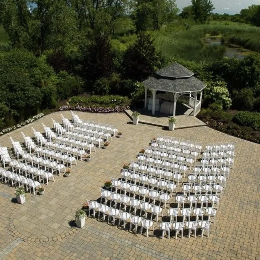 La photo montre une élégante salle de réception en plein air au Plaza Rive Sud, catégorie salle de réception. On y voit un gazebo blanc au centre du fond, entouré de verdure luxuriante. Devant le gazebo, se trouvent plusieurs rangées de chaises blanches parfaitement alignées sur une terrasse pavée, prêtes à accueillir les invités d