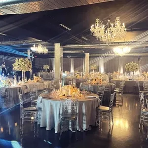 The image showcases a beautifully decorated wedding venue at Plaza Rive Sud, categorized as a wedding venue for Mariage Elegant in Canada. The spacious indoor setting is elegantly adorned with round tables covered in pristine white tablecloths, each accented by exquisite floral centerpieces of white and cream flowers. Crystal chandeliers hang from the ceiling, casting a warm glow over the room and creating a romantic ambiance. The light streams through the venue, highlighting the transparent Chiavari chairs that surround each table. The setting is designed for a sophisticated and memorable wedding reception, capturing the essence of elegance and charm.