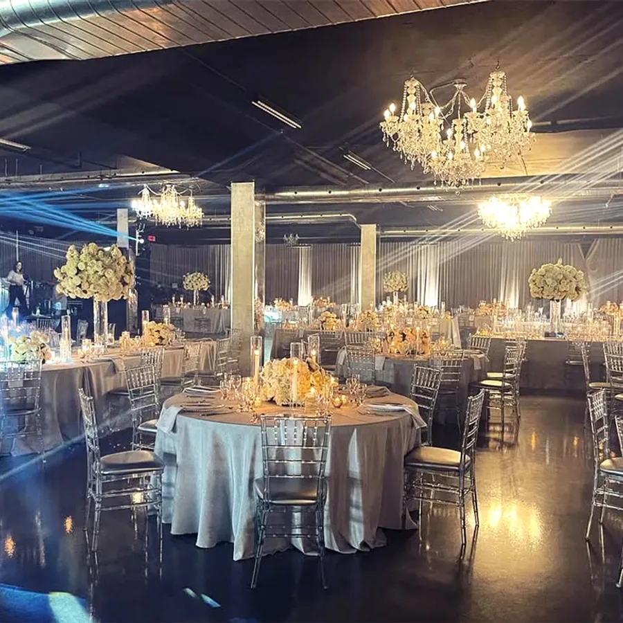 La salle de réception Plaza Rive Sud est magnifiquement décorée pour un mariage élégant. Les tables rondes sont recouvertes de nappes blanches et ornées de centres de table floraux avec des roses blanches. Les chaises en plexiglas transparent ajoutent une touche moderne. De magnifiques lustres en cristal suspendus au plafond diffusent une lumière douce, créant une ambiance romantique. Des projecteurs lumineux accentuent les arrangements floraux et ajoutent une atmosphère festive à l