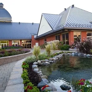 The image showcases Plaza Rive Sud, a stunning wedding venue listed on Mariage Elegant, Canada. The venue features a charming red brick building with a modern, sloped metal roof. A beautifully landscaped garden surrounds a serene pond, complete with lush greenery and colorful flowers, creating an inviting and romantic atmosphere for weddings. The paved walkway and well-maintained hedges add to the elegance of the venue, making it a perfect choice for couples seeking an enchanting outdoor wedding location.