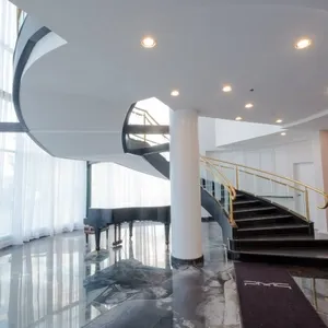The image showcases the elegant interior of Plaza Vogue, a premier wedding venue under the category of Wedding Venues, featured on Mariage Elegant, Canada. It captures a sweeping, modern spiral staircase with a sleek black handrail, accompanied by a polished black grand piano placed near the base. The setting is illuminated by soft, natural light streaming through large, sheer curtain-covered windows, highlighting the glossy marble-like floor. This sophisticated decor reflects an ideal location for hosting luxurious wedding events.