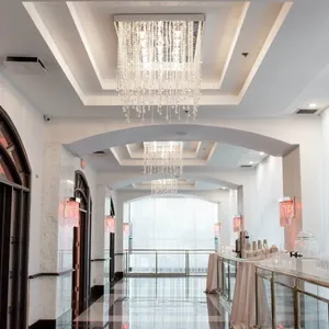 This image showcases a stylish interior view of the wedding venue Plaza Vogue, under the category of Wedding Venues, on Mariage Elegant, Canada. The venue features an elegant hallway with high, arched ceilings and luxurious chandeliers adorned with sparkling crystals that cast a soft glow. The walls are painted in a soothing white and are complemented by dramatic arched windows with dark wood frames. The sleek, polished flooring enhances the sophisticated ambiance. Tables along the side are draped in beige cloths, adding a touch of subtle elegance. This venue exudes a refined and modern aesthetic, ideal for a memorable wedding celebration.