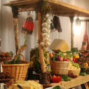 The image showcases an elegantly decorated display at Plaza Vogue, a featured vendor in the Wedding Venues category on Mariage Elegant, Canada. The setting includes a rustic wooden stall adorned with hanging garlic and cured meats, highlighting a rich selection of colorful fruits and vegetables in baskets, alongside a variety of cheese wheels. This visually appealing arrangement creates an inviting and sophisticated atmosphere ideal for a wedding reception, emphasizing the venue's commitment to quality and presentation.