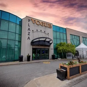 The image shows the exterior of Plaza Vogue, a prominent wedding venue in the Mariage Elegant vendor listing in Canada. The building features a modern design with large, curved glass windows and a stylish entrance canopy. A sign displaying 'VOGUE PLAZA' is prominently positioned above the entrance. The venue is surrounded by a well-maintained area with planters and a small patio, visible on the right. The sky is painted with hues of pink and purple, suggesting a serene and picturesque setting, ideal for weddings.