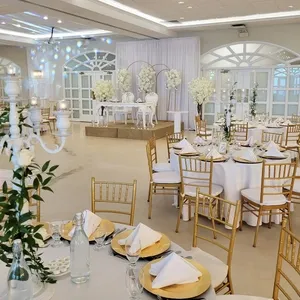 This image showcases the elegant wedding venue Plaza Vogue, categorized under Wedding Venues for Mariage Elegant in Canada. The venue features a beautifully decorated reception hall with round tables draped in white tablecloths, adorned with gold plates and neatly folded white napkins. Each table is surrounded by gold Chiavari chairs, enhancing the luxurious ambiance. The room is elegantly lit, with decorative chandeliers and soft, ambient lighting. In the background, a raised stage is set up with an ornate white backdrop and a floral arrangement, offering a perfect spot for wedding ceremonies or speeches. The overall setting exudes sophistication and charm, ideal for hosting a memorable wedding celebration.