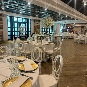 The image showcases the elegant interior of Plaza Vogue, a wedding venue listed under Mariage Elegant, Canada. The room features round tables adorned with white tablecloths and gold charger plates, each set with fine glassware and neatly folded white napkins. Stylish white chairs with intricate designs surround the tables. A centerpiece with white flowers adds a touch of sophistication. The venue's ceiling has a modern design with recessed lighting, and a crystal chandelier can be seen in the corner, enhancing the formal ambiance perfect for a wedding reception.