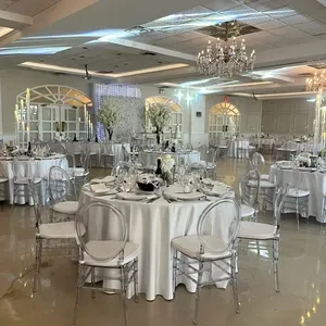 The image shows an elegantly decorated wedding venue offered by Plaza Vogue, categorized under Wedding Venues. The spacious room features multiple round tables draped with pristine white tablecloths and set with fine dinnerware. Transparent, modern chairs surround the tables, complementing the sophisticated décor. Crystal chandeliers hang from the ceiling, adding a touch of luxury and reflecting light across the polished floor. A backdrop of white and pastel flowers can be seen, enhancing the romantic ambiance ideal for a wedding celebration.