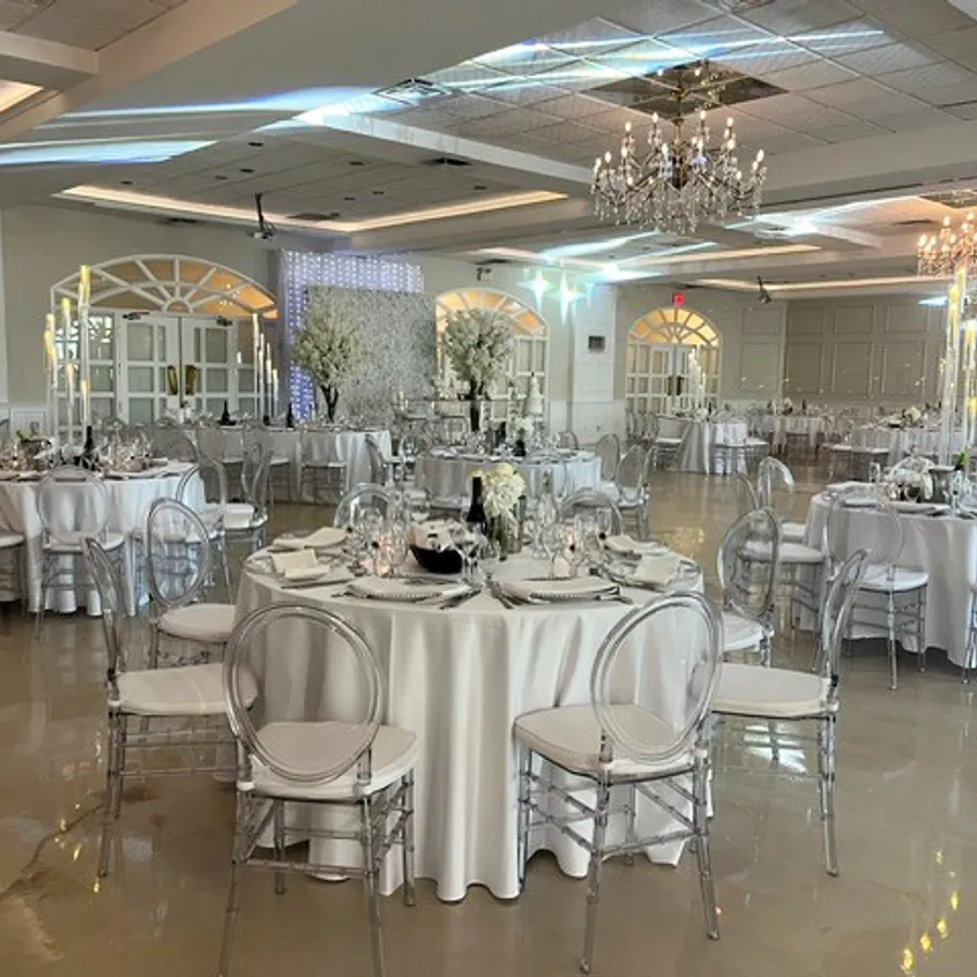 Cette image montre une élégante salle de réception de mariage, fournie par Plaza Vogue, située au Canada. La salle est somptueusement décorée avec des tables rondes recouvertes de nappes blanches, chaque table étant entourée de chaises en acrylique transparent. Au centre, des arrangements floraux blancs ajoutent une touche de sophistication. De magnifiques lustres en cristal pendent du plafond, éclairant la pièce d