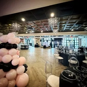 The image showcases an elegantly decorated wedding venue at Plaza Vogue, categorized under wedding venues for Mariage Elegant in Canada. The spacious room features a beautifully arranged seating area with round tables covered in black linens, each adorned with chic table settings and glassware. Overhead, stunning chandeliers hang from a coffered ceiling, casting warm light throughout the space. A large, sophisticated balloon arrangement in shades of pink and black adds a festive touch to the venue. The polished wooden floor and large windows create an inviting ambiance, perfect for hosting a memorable wedding reception.
