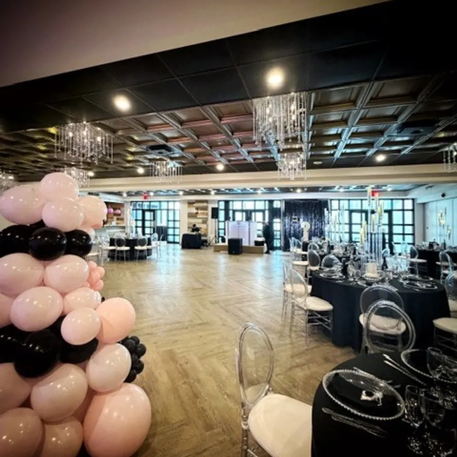 La photo montre une salle élégante de Plaza Vogue, dans la catégorie salle de réception de Mariage Elegant, Canada. On y voit un espace spacieux avec un parquet en bois à chevrons, décoré pour un mariage. Le plafond est orné de lustres en cristal qui ajoutent une touche de glamour. À gauche, il y a une installation artistique faite de ballons roses et noirs, tandis que les tables sont dressées avec des nappes noires, des assiettes en verre et des chaises transparentes, créant une ambiance sophistiquée et moderne.