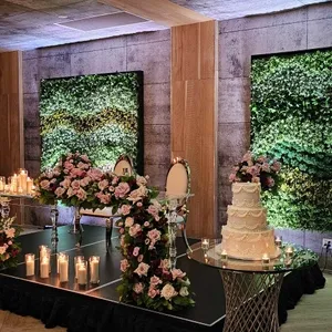 La salle de réception Plaza Volare présente une scène élégante pour un mariage, avec des panneaux en bois au mur et un éclairage suspendu aux ampoules modernes. Un espace surélevé est décoré de chaises sophistiquées et de nombreuses fleurs roses et blanches. Des bougies allumées entourent l'estrade, créant une ambiance chaleureuse. À droite, un gâteau de mariage à plusieurs étages est placé sur une table en métal au design moderne. Les murs sont recouverts de verdure, ajoutant une touche naturelle à la décoration chic de la salle.
